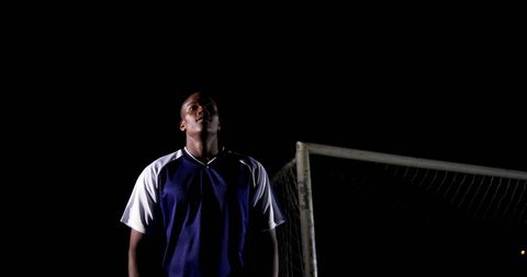 Soccer player stands on the field with a dark background, showcasing his focus as he stares ahead, possibly during a night-time practice or match. The presence of the goalpost in the vicinity suggests he is engaged in the sport competitively. Ideal for use in sporting event promotions, athlete inspiration articles, and advertisements relating to dedication in sports.