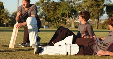 White teen cricketers resting on field wearing pads and holding bat showing team camaraderie