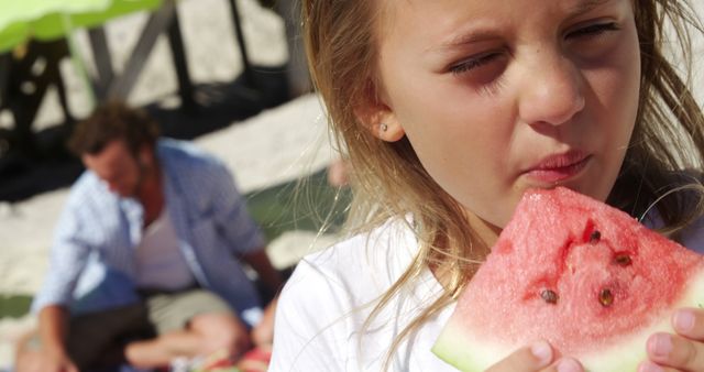 A young Caucasian girl enjoys a slice of watermelon on a sunny beach day, with a man in the background. Her expression suggests she is savoring the sweet, refreshing taste of the fruit during a leisurely outdoor activity.