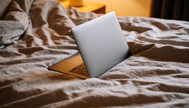 Silver laptop nesting in warmly lit rumpled bed for cozy remote work relaxation