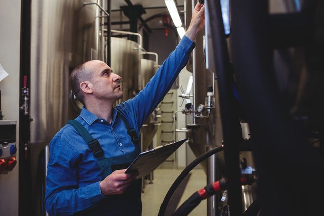 Male manufacturer in a brewery examining machinery with a clipboard. Useful for illustrating industrial processes, quality control, and professional work environments in the brewing industry.