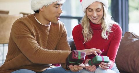 Couple exchanging christmas gifts on sofa wearing santa hats and cozy knit sweaters