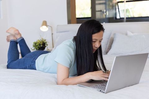 Mature asian woman lying on bed using laptop in sunlit bedroom for remote work