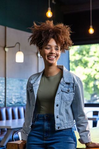 Smiling african american woman standing in cozy cafe wearing denim jacket and jeans