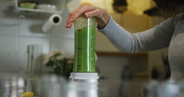 Woman blending green smoothie in kitchen using modern appliances. Represents healthy lifestyle, fresh ingredients, and home cooking.Commercial use includes ads for kitchen appliances, health blogs, food-related websites, and cooking tutorials.