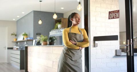 African american barista leaning at open cafe door wearing apron, modern cozy coffee shop