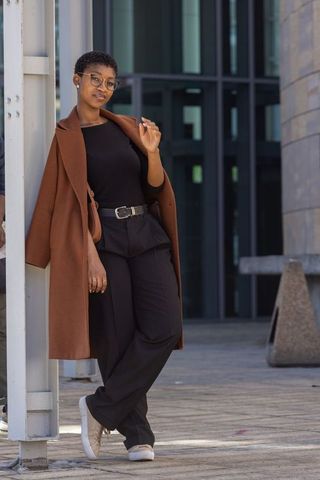 African woman leaning on column wearing brown coat and glasses in modern urban plaza
