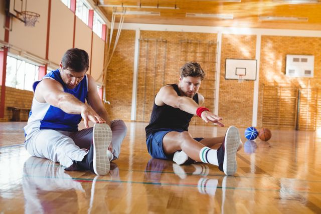 Two male friends are stretching on a basketball court. They are sitting on the floor, reaching towards their feet. The gym setting includes basketballs and a hoop in the background. This image is ideal for promoting fitness, teamwork, and healthy lifestyles. It can be used in sports-related articles, fitness blogs, or advertisements for gym memberships and athletic programs.