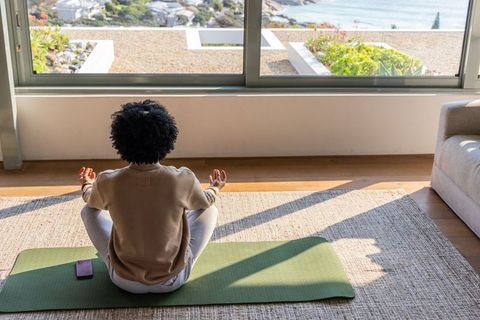 African american woman meditating on yoga mat in sunny coastal living room with ocean view