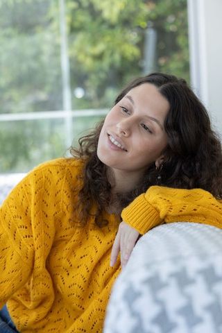 Young woman reclining on sofa wearing mustard sweater gazing through sunlit window