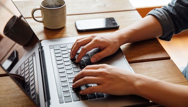 Hands typing on laptop keyboard at wooden desk with coffee mug and smartphone, remote work