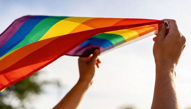 Hands raising sunlit rainbow pride flag overhead waving in park celebrating lgbtq+ pride