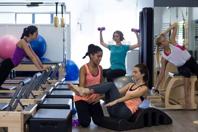 Female trainer assisting woman with stretching exercise on arc barrel in gym. Other women are engaged in various fitness activities in the background. Ideal for use in fitness blogs, gym advertisements, pilates class promotions, and health and wellness articles.