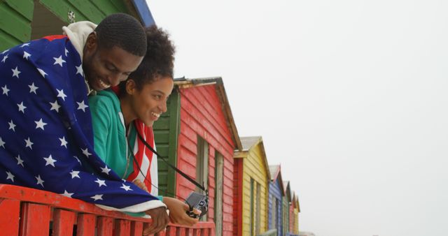 Couple wrapped in USA flag smiling outdoors at vibrant beach houses. Perfect for themes of travel, romance, celebration, patriotism, and seaside journeys.