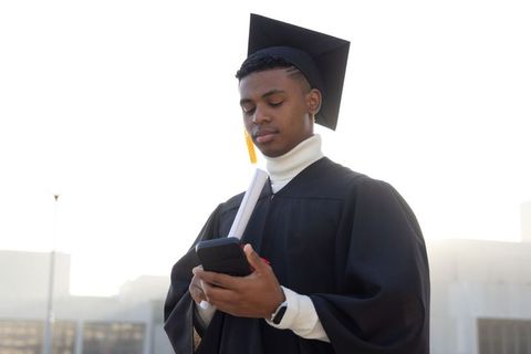 Young african american graduate wearing cap and gown checking smartphone holding diploma