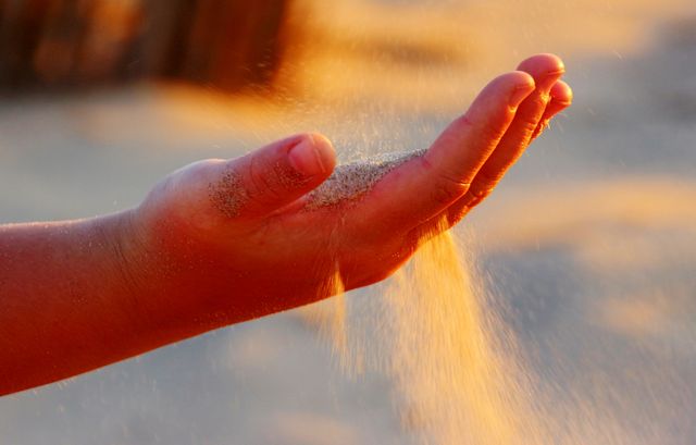 Close-up of a hand holding and releasing sand, with the golden light of sunrise in the background. This stock photo can be used for themes related to vacation, relaxation, nature, time passing, environmental conservation, summer activities, and tranquility.