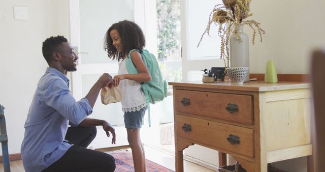 Happy african american father giving smiling daughter packed lunch before she leaves for school. Fatherhood, childhood, care, togetherness and domestic life.