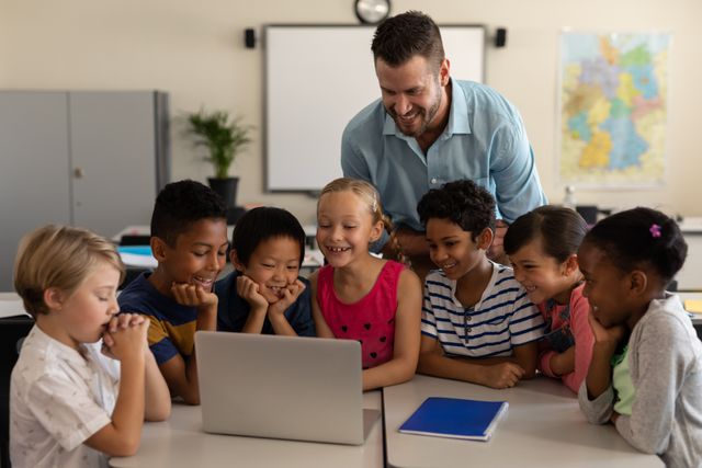 Male teacher engaging a group of diverse elementary school students with a laptop in a classroom. Ideal for educational content, technology in education, classroom activities, and promoting interactive learning environments.