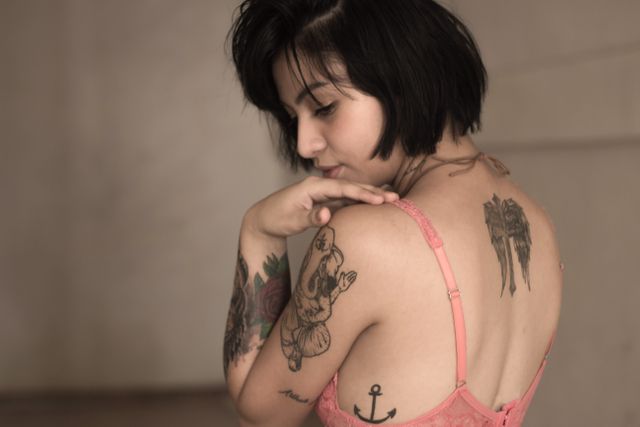 Young woman with several tattoos on her back and arms, wearing pink lingerie. Presenting a back view with emphasis on inked artwork including an anchor and a scholor tattoo. Ideal for use in body art promotions, tattoo studio advertisements, fashion editorials, and self-expression narratives.