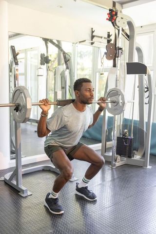 African american man performing loaded back squat in modern gym with mirror and barbell