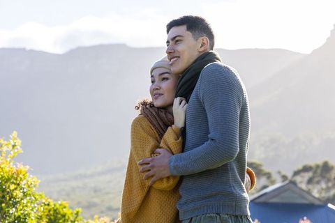 Asian couple embracing on sunlit hillside looking toward horizon wearing knit sweaters