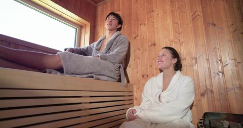 Mixed-race couple relaxing in wooden sauna wearing bathrobes by daylight window urban spa