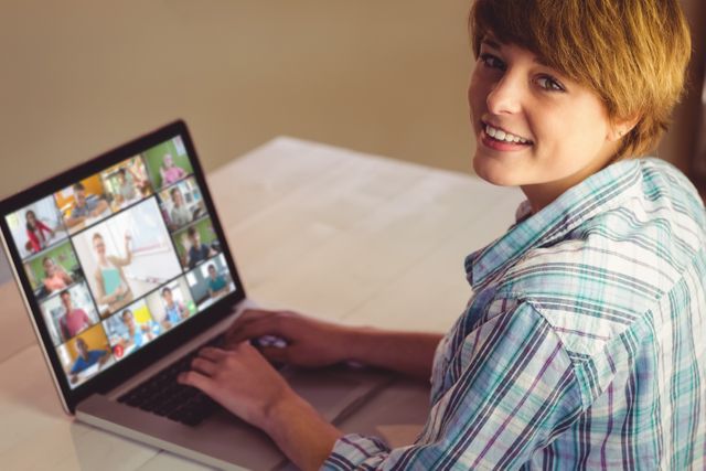 Smiling young woman using laptop in home office for online video conference with multiple participants. Can be used for concepts related to remote work, virtual meetings, online collaboration, digital communication, home workspaces, and modern technology. Ideal for articles, blog posts, and advertisements promoting remote working and digital communication platforms.
