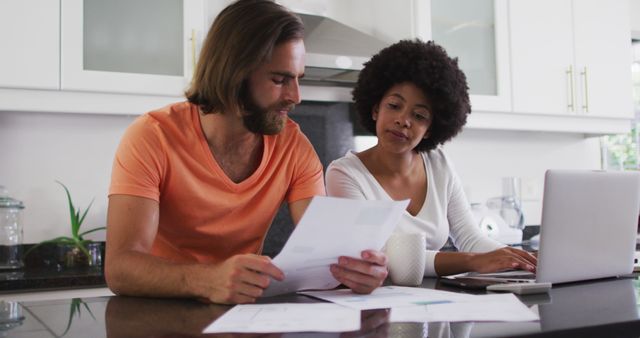 Multiethnic couple at kitchen counter reviewing financial documents with laptop open. Ideal for content on financial planning, household budgeting, teamwork in relationships, or home office setup. Perfect for articles about managing finances as a couple or paying bills together.