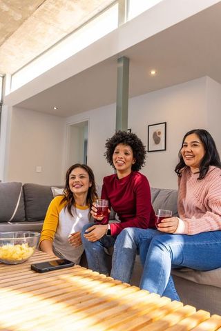 Diverse women laughing and enjoying drinks on modern gray sofa in bright living room