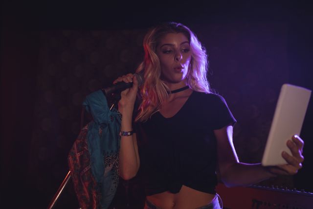 This image captures a female singer taking a selfie with a digital tablet while performing in a nightclub. Ideal for use in articles or advertisements related to nightlife, entertainment, music events, social media, and modern technology. It can also be used for promoting concerts, music venues, or showcasing the blend of technology and live performances.