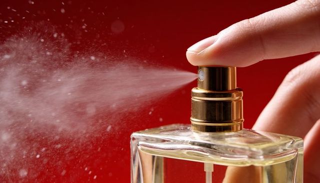 Closeup fingertip pressing gold atomizer spraying perfume mist over rich red background