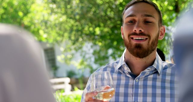 Man holding wine glass, smiling, enjoying casual conversation at outdoor social event. Could be used for promotions involving social gatherings, lifestyle blogs, or advertising casual wear.