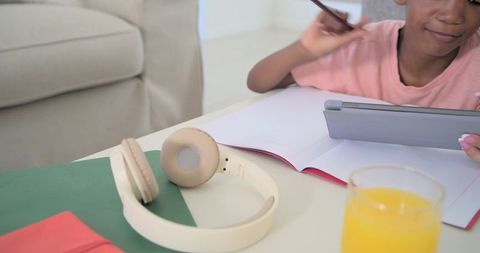 Teen using tablet and stylus studying at home with headphones, notebook and orange juice