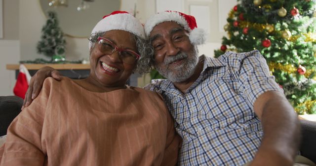 Senior couple wearing Santa hats and sitting on couch in living room, smiling and enjoying Christmas together. Ideal for holiday greeting cards, advertisements promoting senior living, retirement communities, family-oriented marketing, and festive blog posts.
