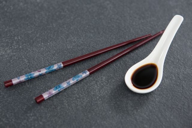 Close-up image of traditional chopsticks and a spoon filled with soy sauce on a stone table. Ideal for use in articles or advertisements related to Asian cuisine, dining, restaurants, or culinary traditions. Perfect for food blogs, recipe websites, and cultural content.