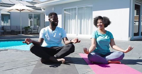 Couple meditating by pool on yoga mats in backyard smiling during wellness routine together