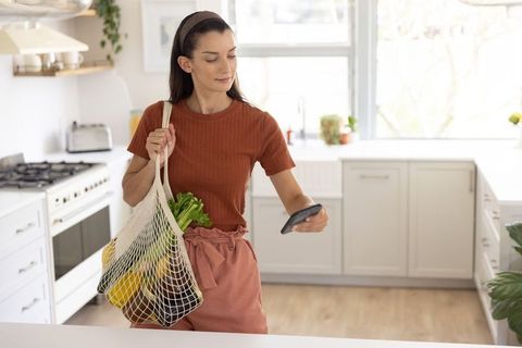 Woman holding reusable mesh grocery bag checking phone in modern sunlit kitchen