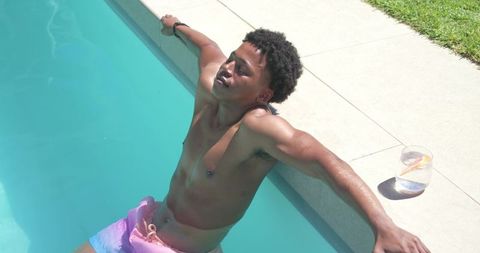 African american man in 20s relaxing at sunlit backyard pool in pastel swim trunks