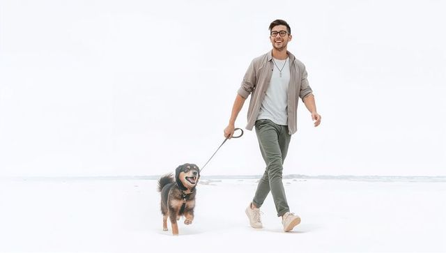 Smiling young man walking happy dog on minimal beach with retractable leash and casual style