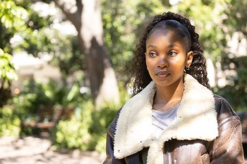 African american woman wearing brown shearling jacket standing in sunlit park thoughtful portrait