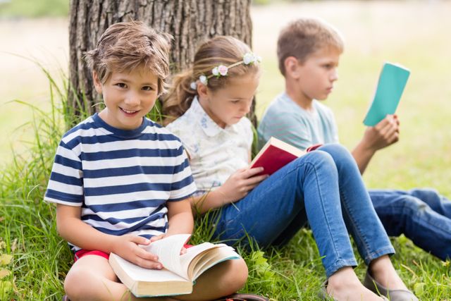 Children sitting under a tree in a park, reading books on a sunny day. Ideal for educational content, summer activities, childhood learning, outdoor leisure, and promoting reading habits among kids.