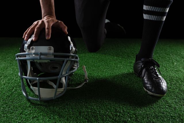 This image captures a close-up of an American football player resting his hand on a helmet placed on artificial grass. Ideal for use in sports-related articles, advertisements for athletic gear, or promotional materials for football events. The focus on the helmet and the player's hand conveys themes of preparation, readiness, and the intensity of the sport.
