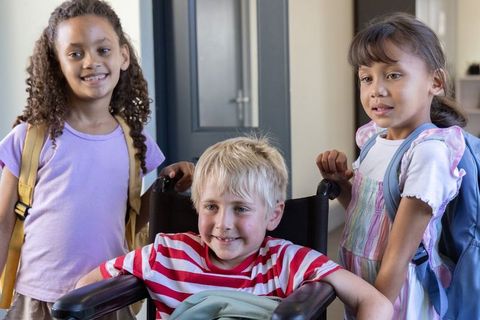Smiling children welcoming classmate in wheelchair at school entrance with backpacks