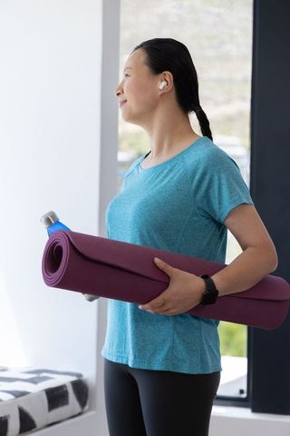 Mature african american woman holding purple yoga mat and bottle, wearing earbuds by window