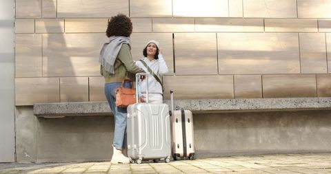 Two women chatting at urban plaza with rolling luggage, stylish bags and winter layers