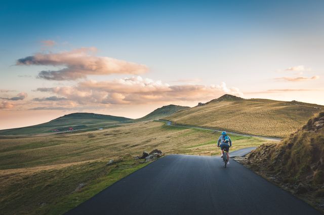 Cyclist biking along picturesque mountain road during sunset, showcasing a serene natural landscape and a sense of adventure. Ideal for use in promotions related to outdoor activities, travel, fitness, wellness, and adventure tourism.