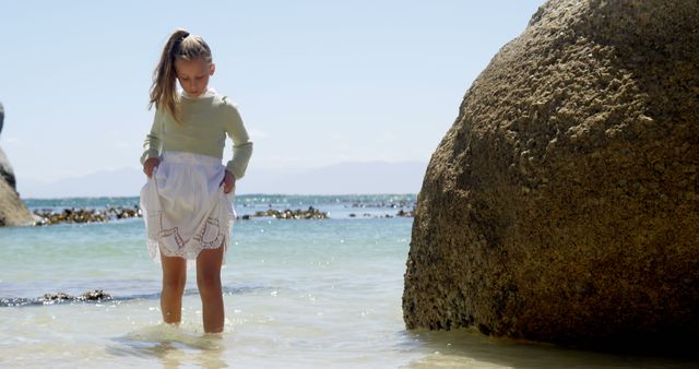 Young girl is playing in ankle-deep sea water near a rocky beach on a sunny day. The clear blue water and large rocks in the background create a serene and peaceful scene. Ideal for use in advertisements, travel blogs, family vacation brochures, and educational materials about marine environments.