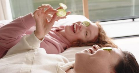 Diverse women relaxing with cucumber eye slices, laughing and enjoying cozy home spa