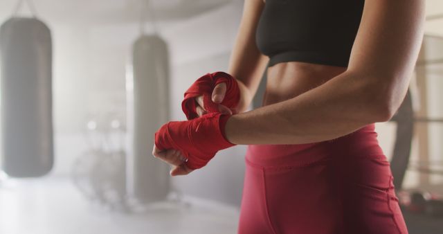Female athlete wrapping her hands in red bandages before a boxing workout at a gym. This image is ideal for illustrating fitness routines, boxing training, sports gear, and promoting an active and healthy lifestyle.
