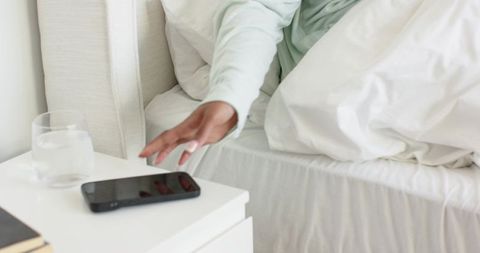 African american woman reaching for smartphone on minimalist bedside table in soft morning light